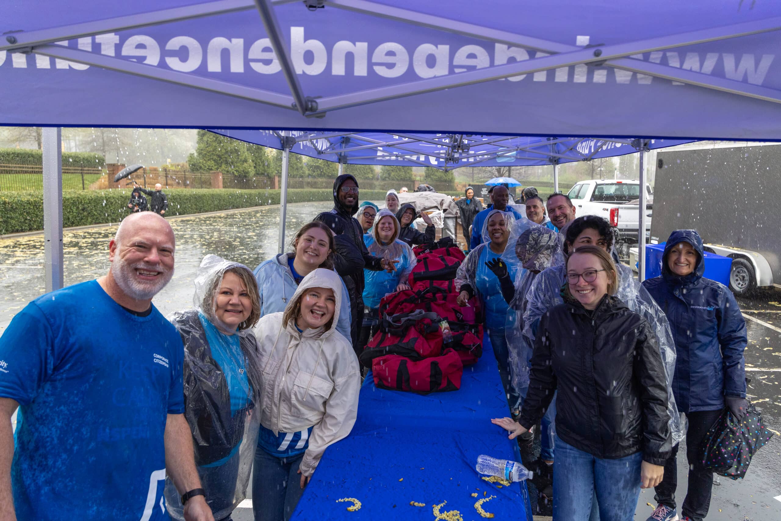 A diverse group of smiling people wearing rain gear under a blue tent during a heavy rain shower, with red duffel bags on a blue table.