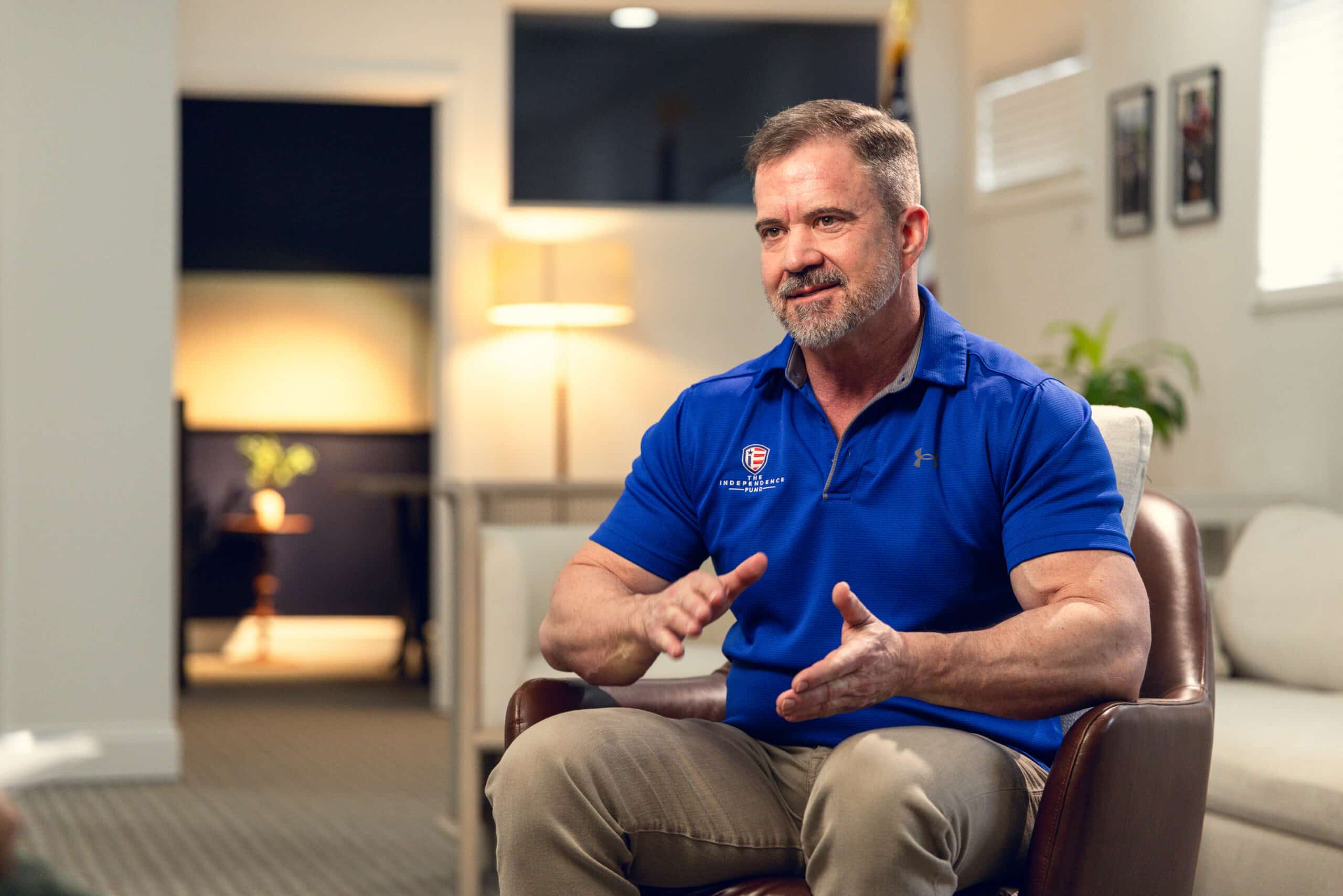 A man with a beard and short hair, wearing a blue polo shirt and khaki pants, sits in a brown leather chair, gesturing with his hands as he speaks.