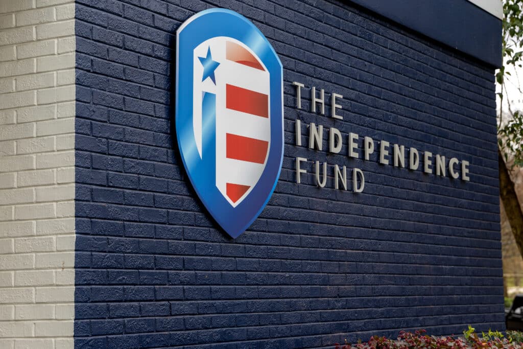 The Independence Fund sign on a dark blue brick wall. The sign has a shield logo with a blue star and red and white stripes, next to the white text 'THE INDEPENDENCE FUND'.