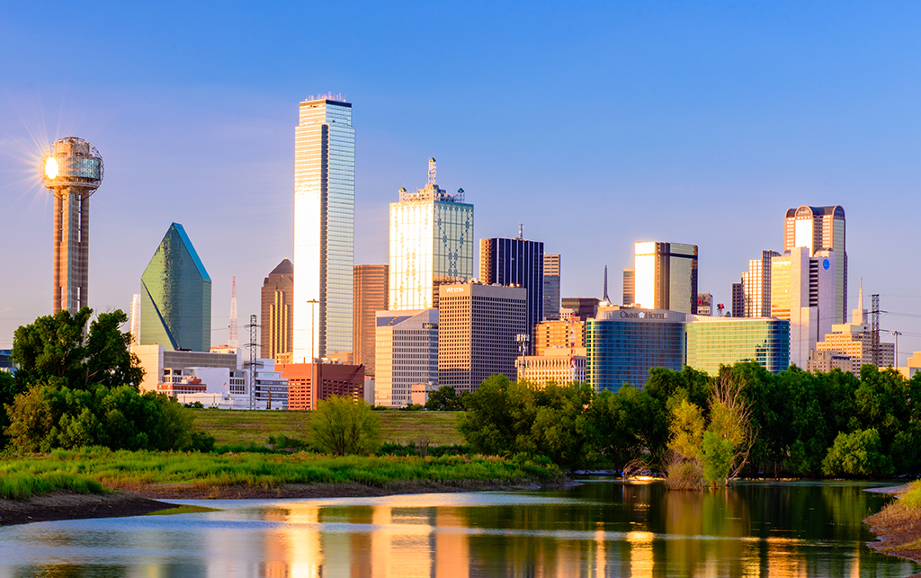 The Dallas, Texas skyline at sunset, featuring Reunion Tower and numerous skyscrapers, reflected in a calm body of water with green trees along the banks.