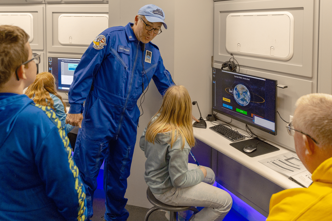 A man in a blue flight suit with a nametag "PAT CRASSE" teaches a young girl at a computer displaying Earth and orbital paths, with other children observing.