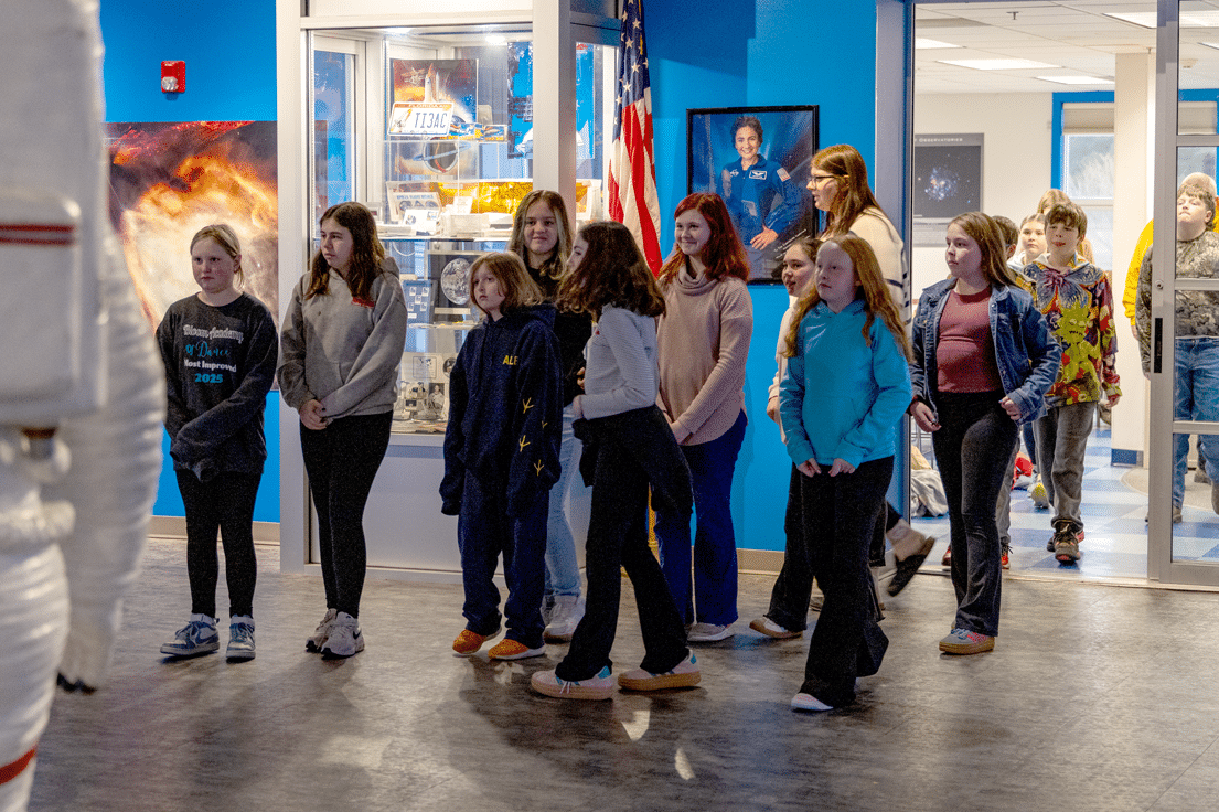 A group of children and young adults observe space exhibits in a museum, with an astronaut suit, display cases, and an American flag visible.