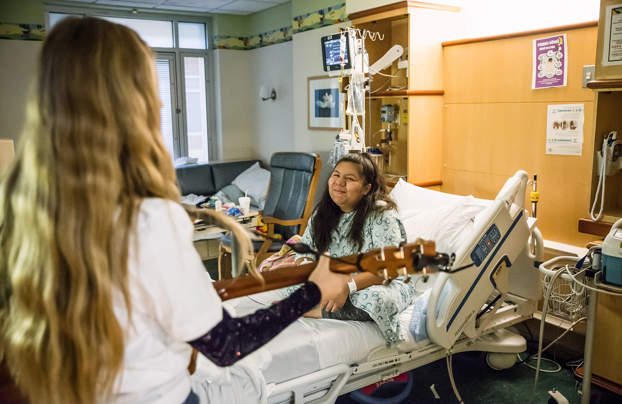 A young woman with long hair plays an acoustic guitar for a smiling patient sitting up in a hospital bed.