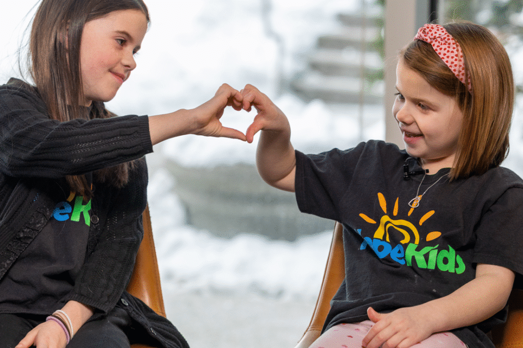 Two young girls, wearing HopeKids t-shirts, form a heart shape with their hands while smiling at each other.