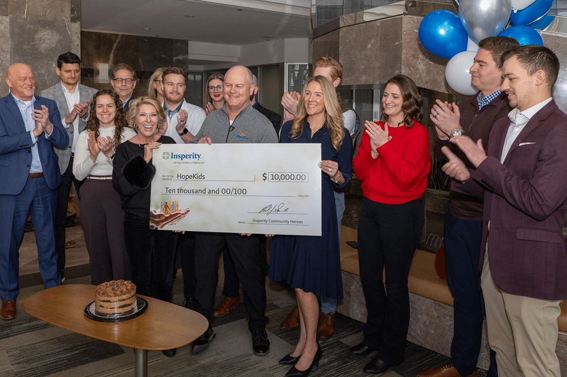 A group of people, including men and women, are gathered around a large check from Insperity to HopeKids for $10,000, clapping and smiling. A chocolate cake is on a table in the foreground.