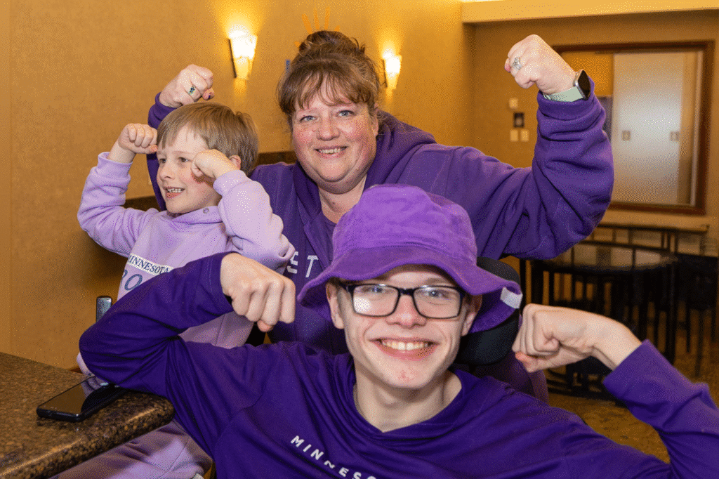 Three people, an adult woman, a young boy, and a teenage boy, all wearing purple clothing, smile and flex their biceps at the camera.