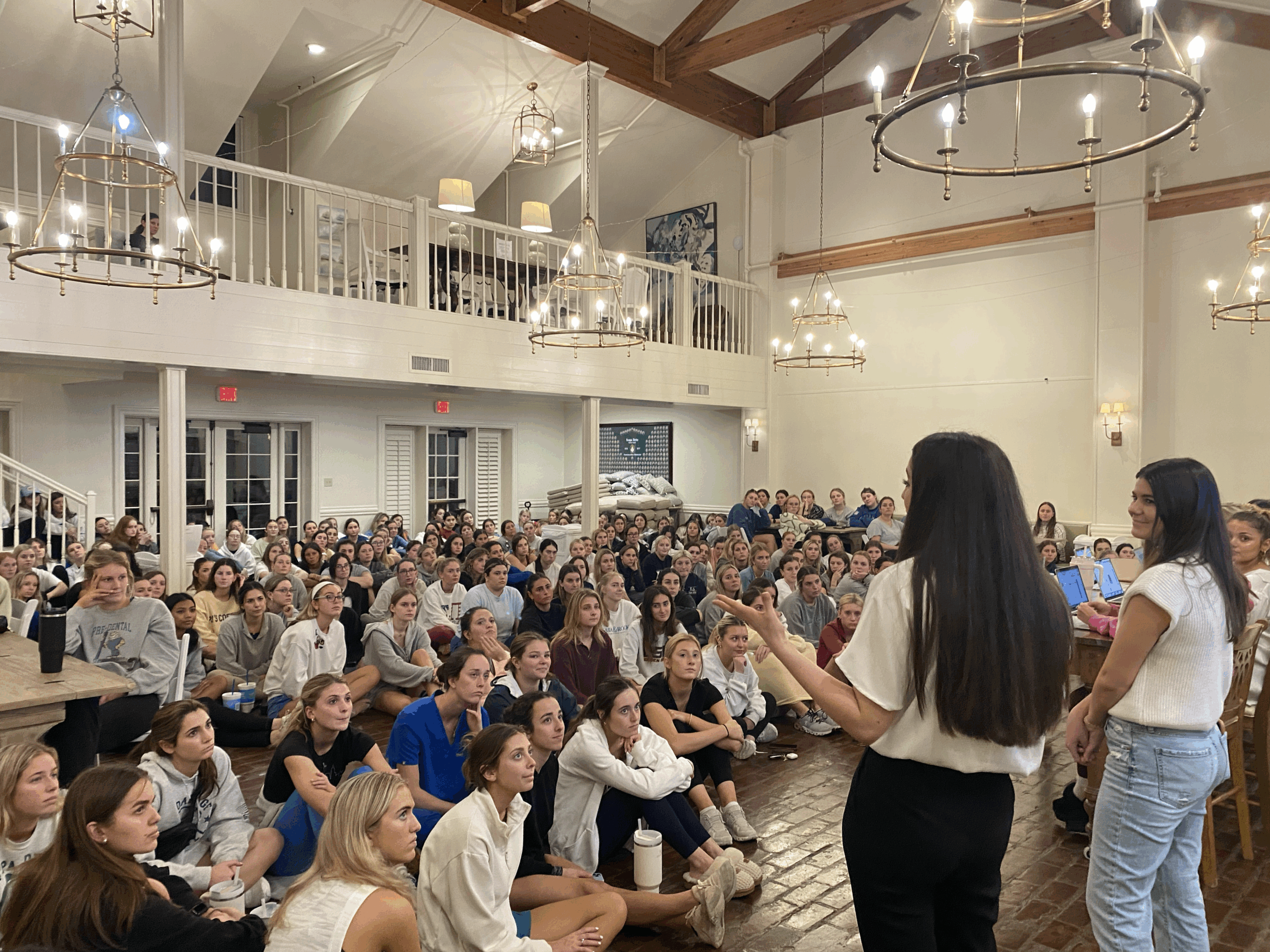 A large group of young women seated on the floor listening intently to two speakers in a spacious hall with high ceilings, chandeliers, and a mezzanine.