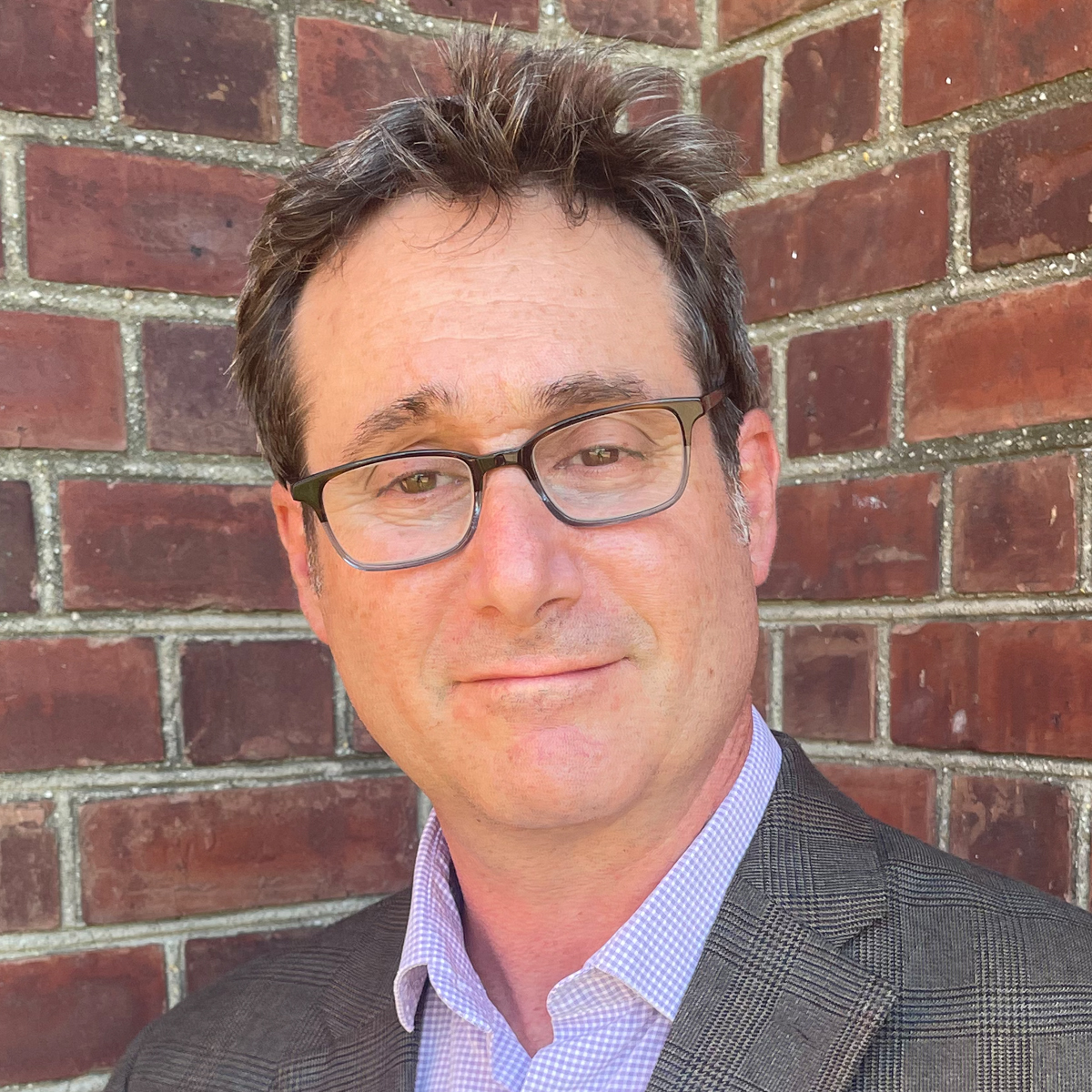 Headshot of a man with glasses and graying hair, wearing a blazer and a checkered shirt, smiling against a red brick wall.