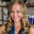 Headshot of a smiling woman with long blonde wavy hair and blue-green eyes, wearing a denim top and silver necklace, against a blurred background of shelves with products.
