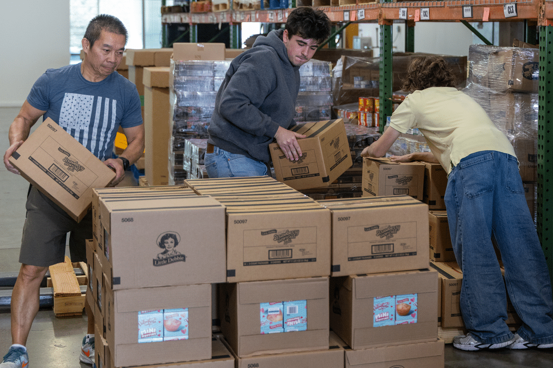 Three people, two men and one person seen from behind, organize stacks of cardboard boxes labeled 'Little Debbie' and 'Strawberry Shortcake Rolls' in a warehouse.