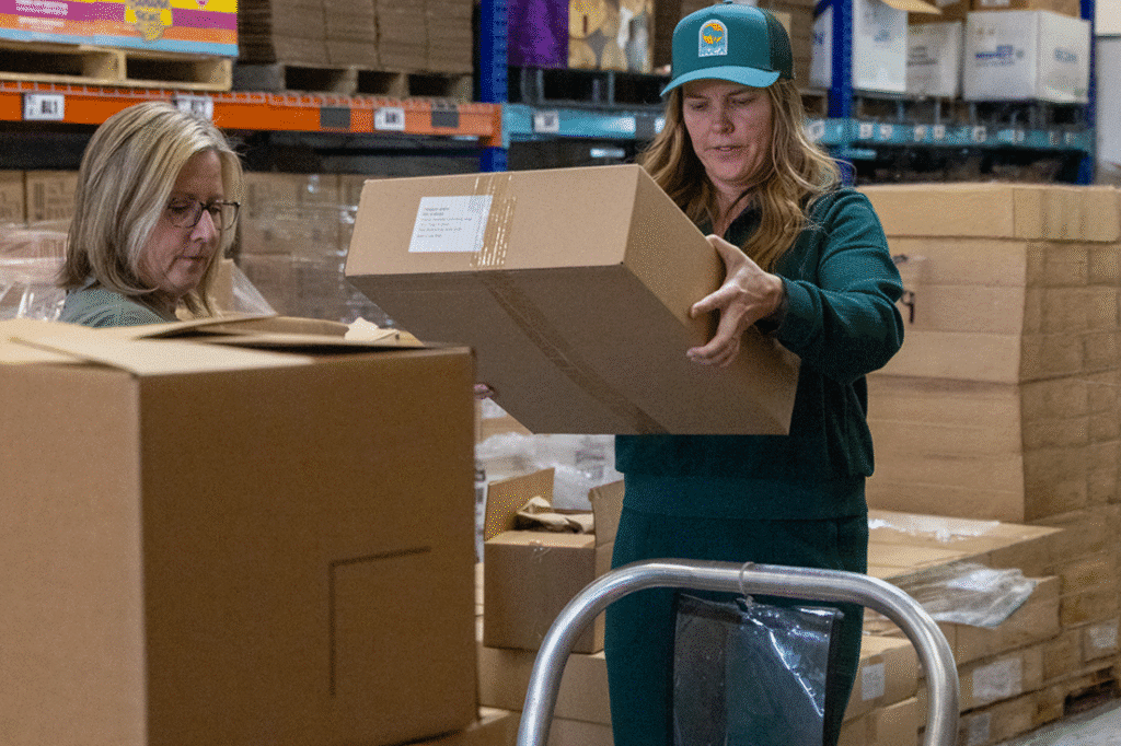 Two women in a warehouse, one handling a cardboard box, surrounded by stacked boxes and shelves.