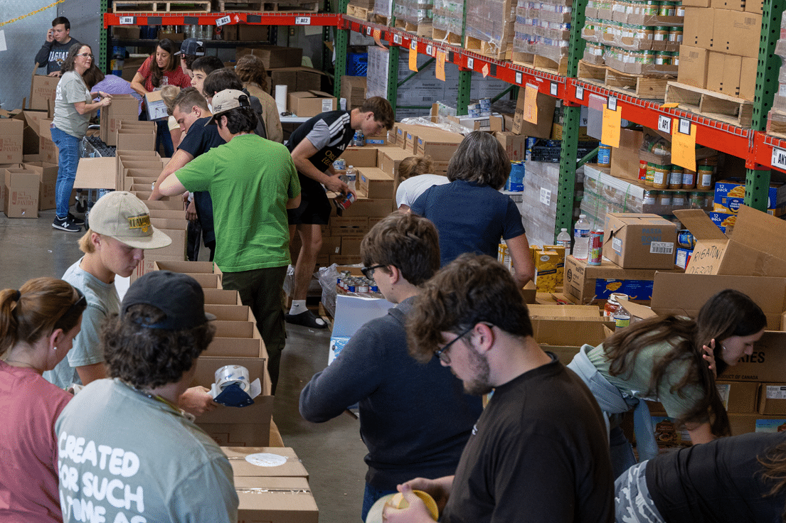 A group of volunteers, mostly young adults, packing food items into cardboard boxes in a large warehouse filled with shelves of packaged goods.