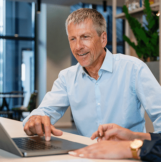 A smiling man points at a laptop trackpad while another person's hands are visible, wearing a gold watch.