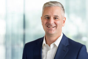 Headshot of a smiling man with graying hair, wearing a navy suit and white shirt, against a blurred office background.
