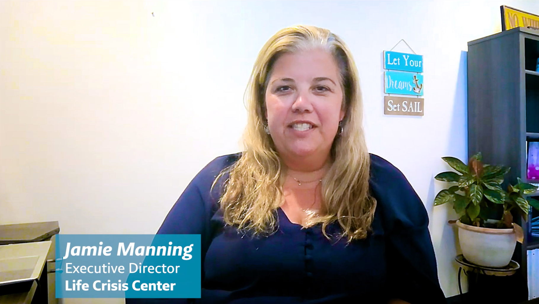 Jamie Manning, Executive Director of Life Crisis Center, smiling in a video call. She has blonde hair and is wearing a dark blue top. In the background, there's a decorative sign that says 'Let Your Dreams Set SAIL' and a potted plant.