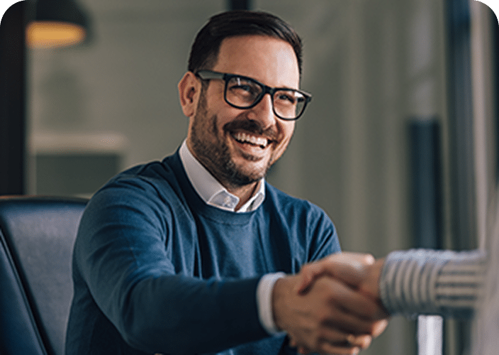 A smiling man in glasses and a blue sweater shaking hands with another person in an office setting.