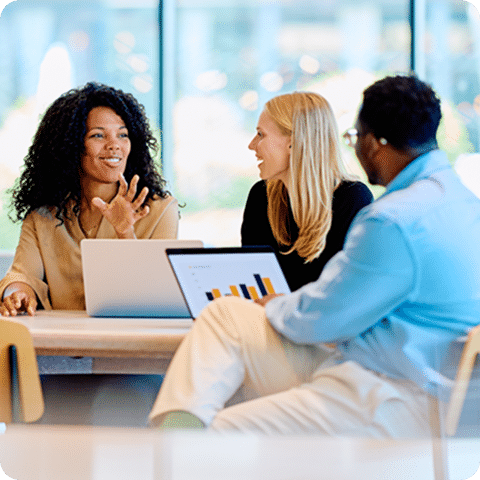 Three people, two women and one man, sit at a table. One woman gestures while speaking, another listens, and the man views a bar chart on a tablet.