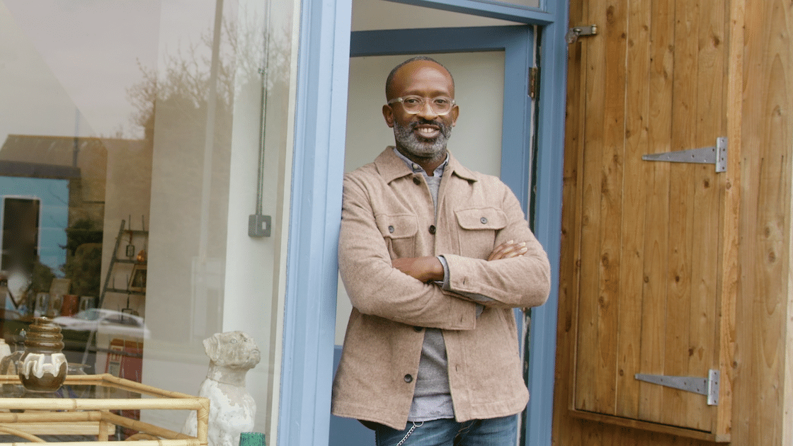 A smiling man with a salt-and-pepper beard and glasses stands in a doorway, arms crossed, wearing a tan shacket. A wooden door is to his right, and a shop window reflecting various items is to his left.