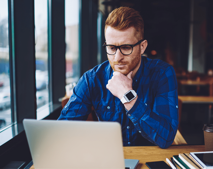A man with red hair and glasses, wearing a blue plaid shirt, looks at a laptop screen with his chin resting on his hand. He wears a white smartwatch. A coffee cup and notebooks are on the table.