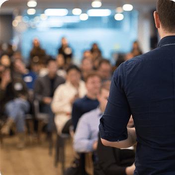 A speaker seen from behind addressing a seated audience in a conference or lecture hall.
