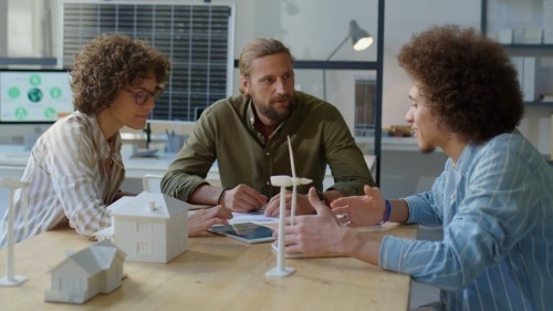 Three people, two men and one woman, sit around a table with 3D models of houses and wind turbines, discussing sustainable design.