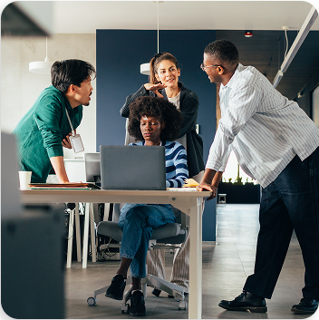 Four diverse professionals, three standing and one seated, collaborate around a laptop in a modern office. The seated Black woman focuses on the screen while her colleagues lean in, discussing.