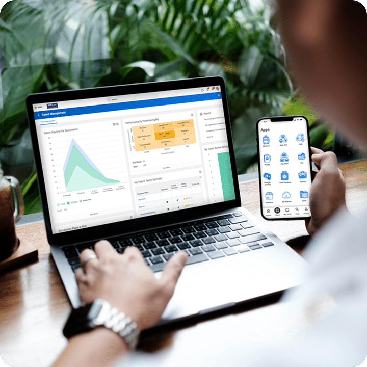 A person uses a laptop displaying a talent management dashboard and holds a smartphone showing an app screen, with green foliage visible outside a window.