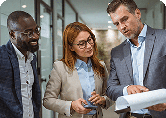 Three business professionals, two men and one woman, reviewing documents on a clipboard in an office hallway.