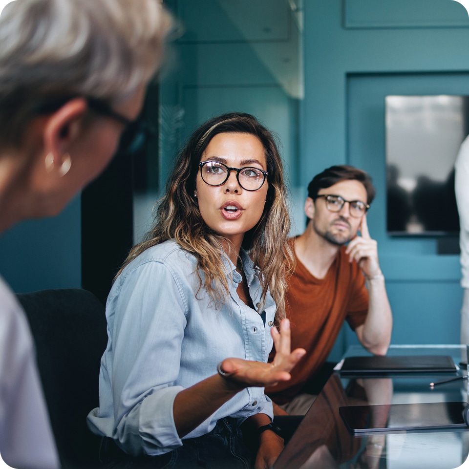 Three people in an office meeting. A woman in glasses speaks, gesturing with her hand, while a man in glasses listens thoughtfully. Part of another person is visible on the left.