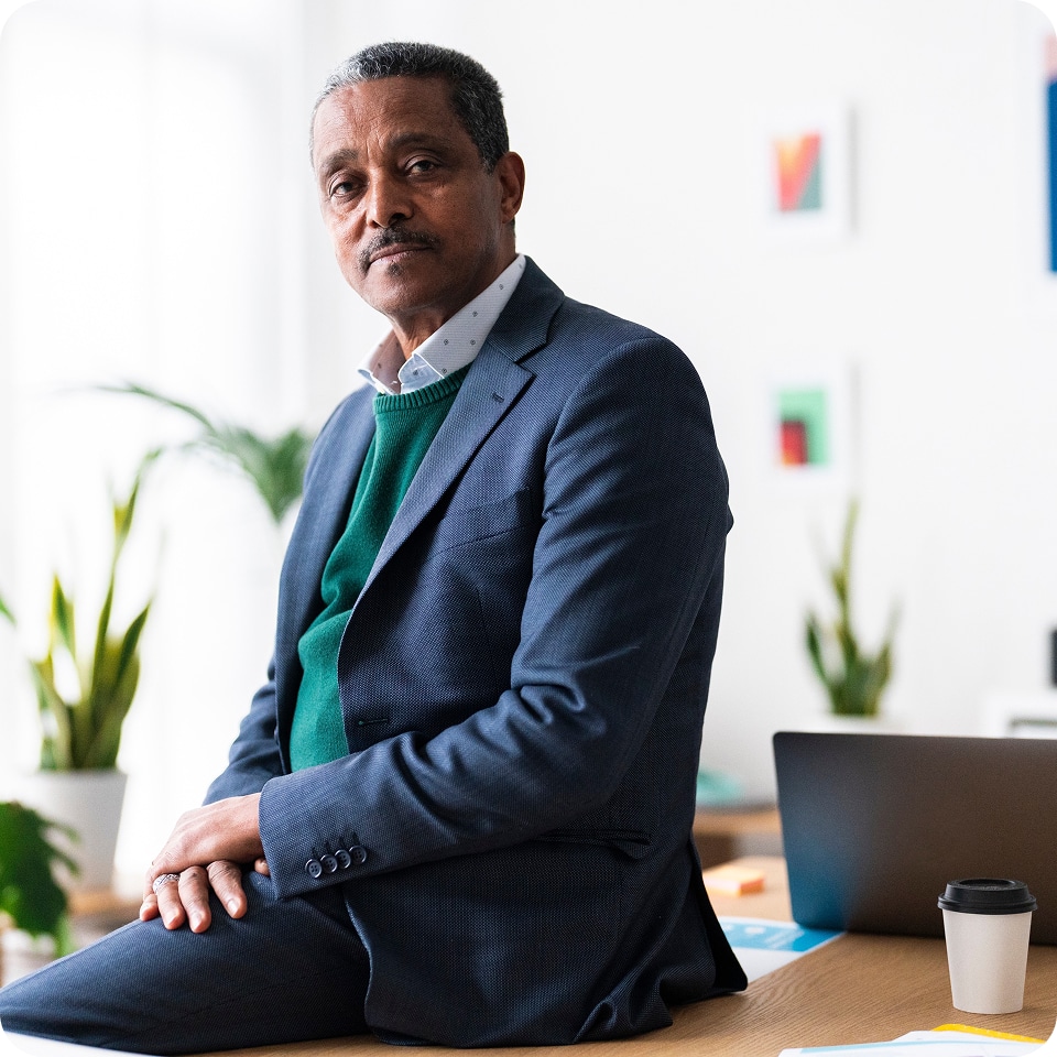 A man in a blue suit and green sweater sits on a desk, looking at the camera. A laptop, coffee cup, and plants are in the background.