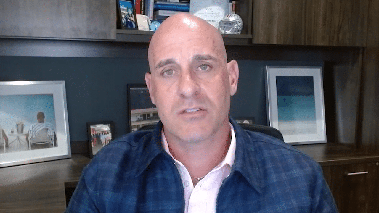 A bald man in a blue plaid jacket and pink shirt looks forward, with an office background featuring framed pictures and shelves.
