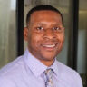 Headshot of a smiling Black man in a light purple shirt and patterned tie.