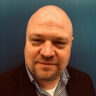 Headshot of a bald man with a beard, wearing a black jacket and patterned shirt, against a blue wall.