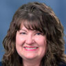 Headshot of a smiling woman with dark brown, wavy hair and bangs, wearing a dark top against a blue-grey background.