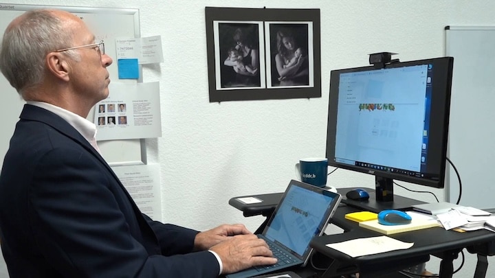 A man with grey hair and glasses, wearing a blazer, works at a standing desk with a laptop and external monitor.
